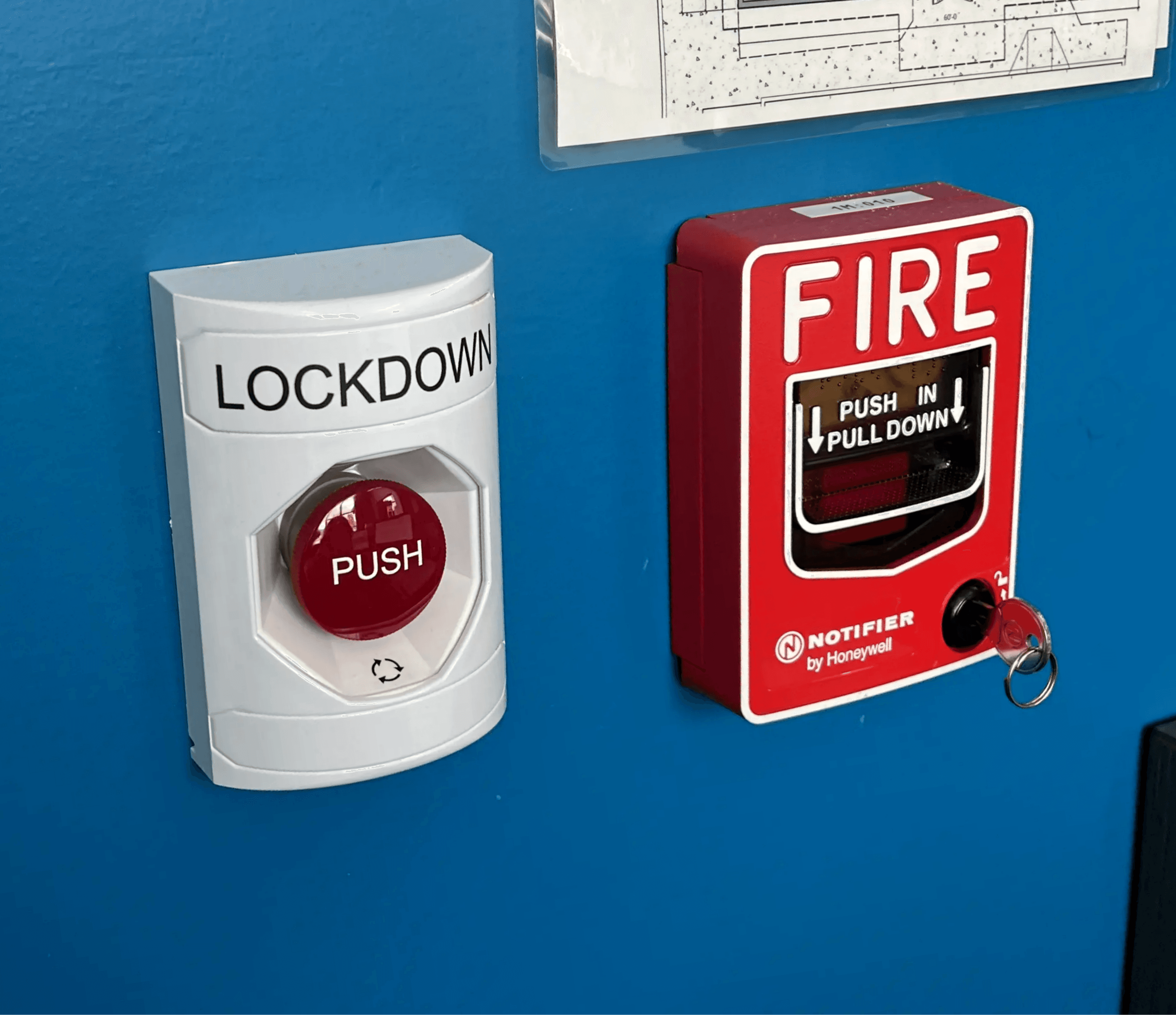 Emergency lockdown and fire alarm manual pull stations mounted on a bright blue wall next to a building's fire escape plan, exemplifying Axe Fire & Security's comprehensive installation services for safety systems.